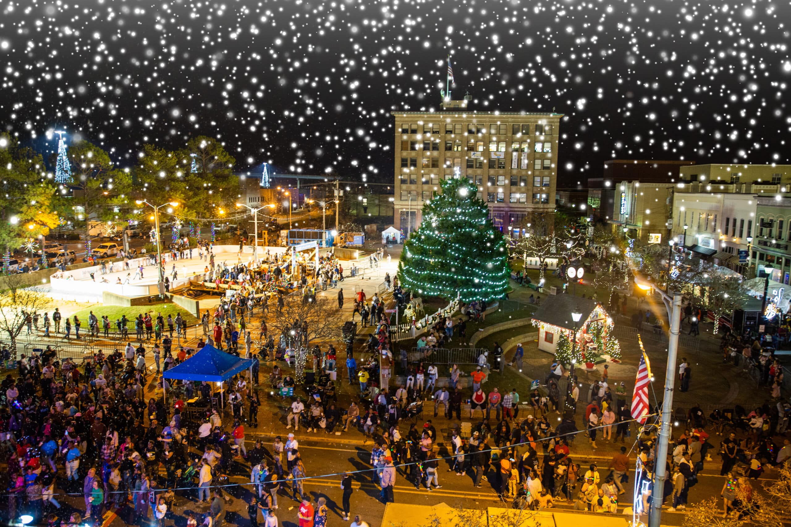An aerial view of Alex Winter Fête bustling with crowds exploring food and art/retail vendors. Illuminated vendor booths surround a decorated central Christmas tree, while festival attendees enjoy food stalls, holiday-themed activities, and an ice skating rink under twinkling lights and falling snow.